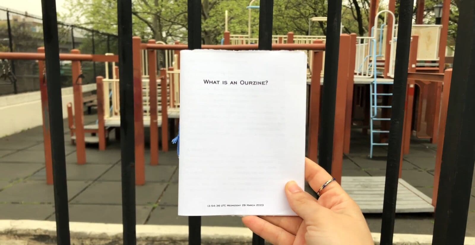 A hand holding up a small booklet titled What is an Ourzine? in front of a playground